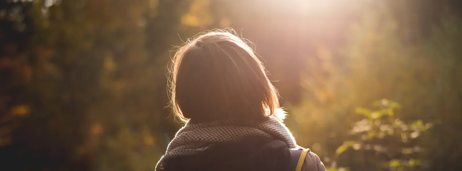 Person with shoulder-length hair wearing a scarf, looking towards sunlight in a forested area during autumn.