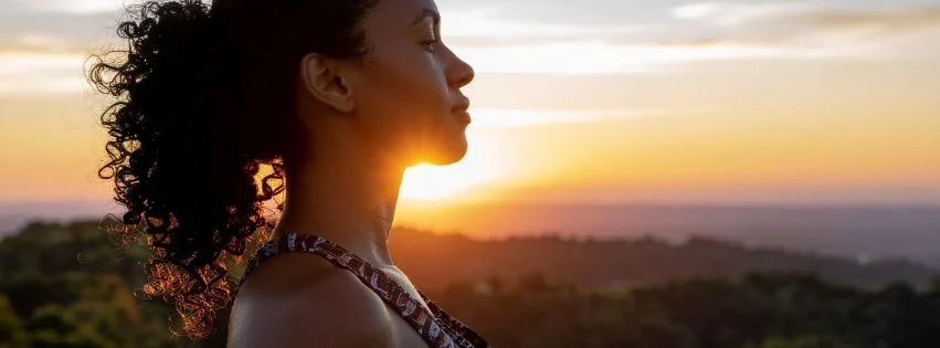 Woman standing outdoors at sunset, looking toward the horizon, representing wellbeing, hope, and a renewed sense of health and balance.
