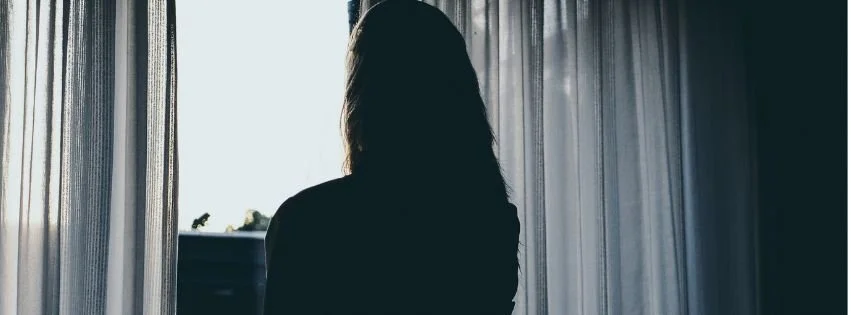 Woman standing by a window in a dimly lit room, representing the emotional changes and social withdrawal that can occur with PMS.