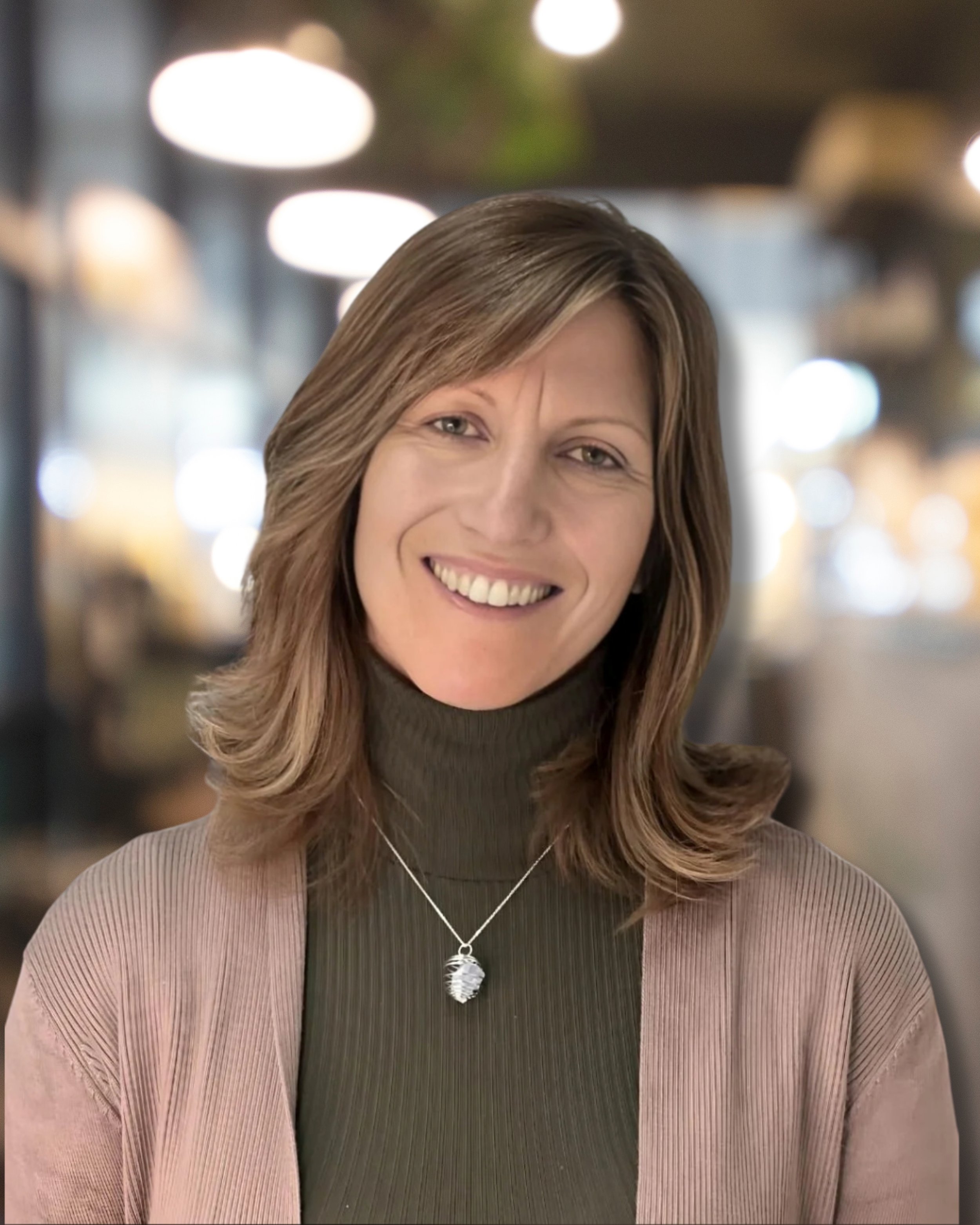 Close-up of a woman with light brown, wavy hair smiling, in a setting with blurred bright lights in the background. She is wearing a black turtleneck sweater, a pink cardigan, and a silver necklace with a shell pendant.