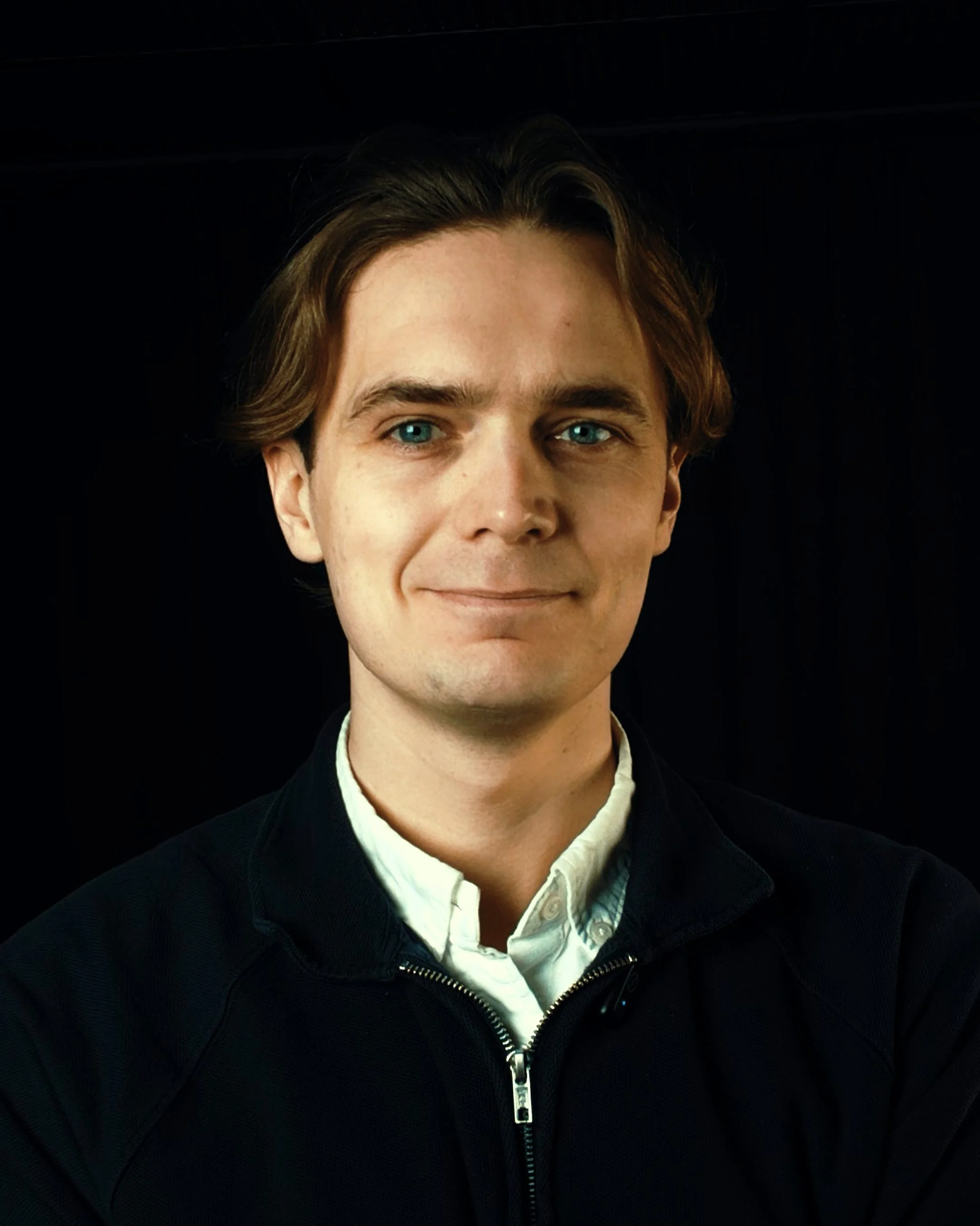 Young man with blue eyes and light brown hair smiling in front of a dark background, dressed in a white collared shirt and a black zip-up jacket.