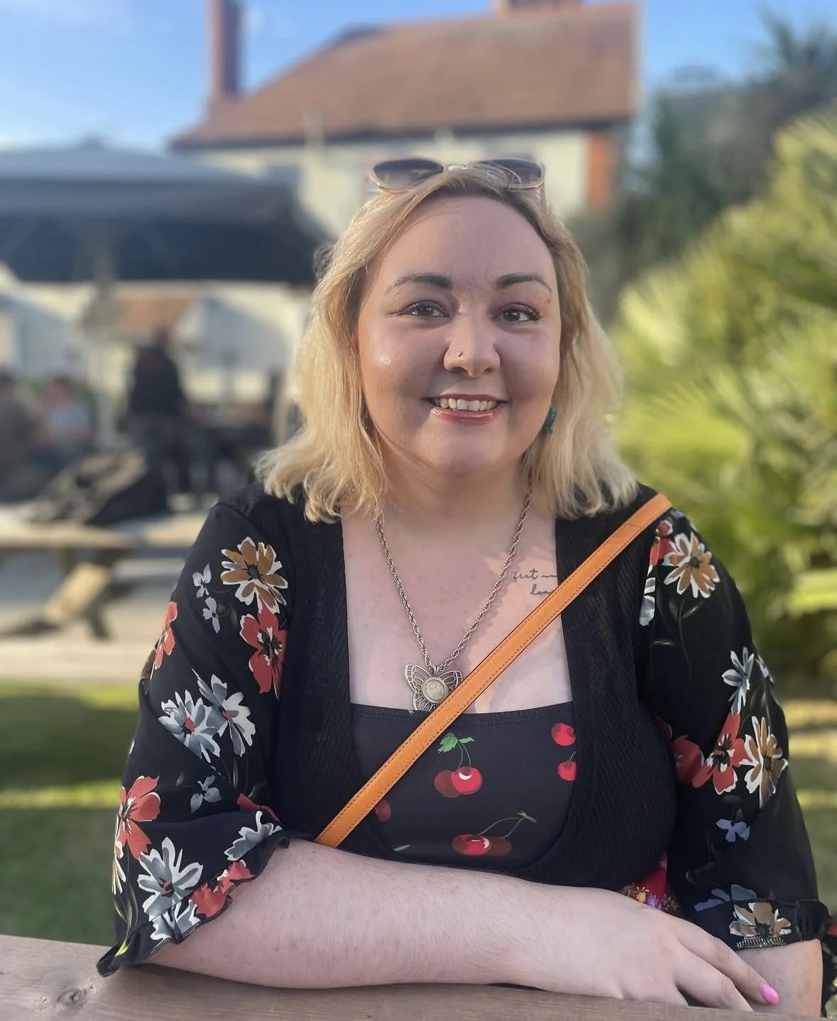 A smiling woman with blonde hair and sunglasses on her head sitting outdoors at a wooden table, wearing a black dress with a cherry pattern and a floral shawl, with a background of people, picnic tables, and a house with a red roof.