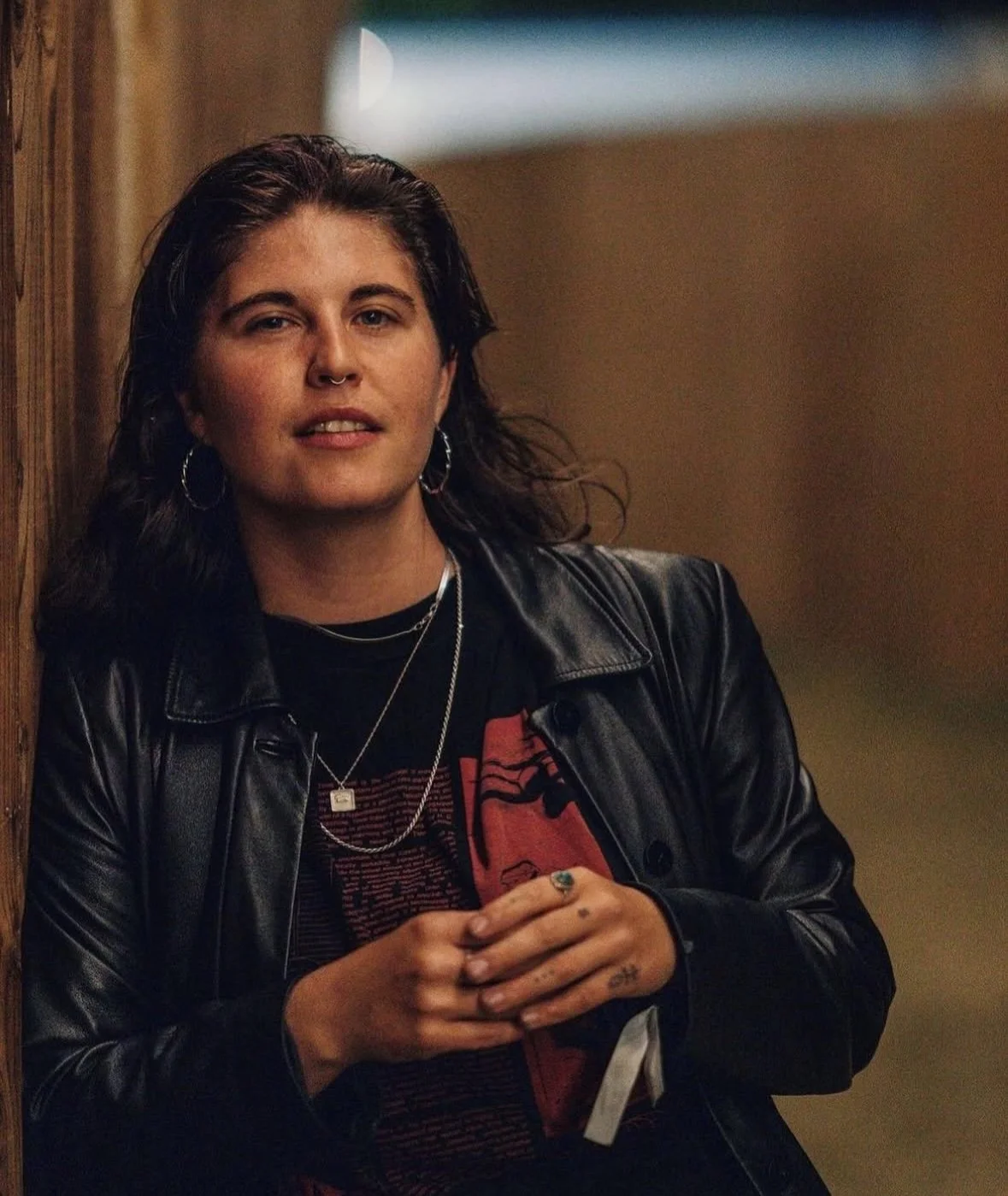 A young woman with dark hair, wearing a black leather jacket, hoop earrings, layered necklaces, and a graphic T-shirt, standing indoors against a brown wall with a window in the background.