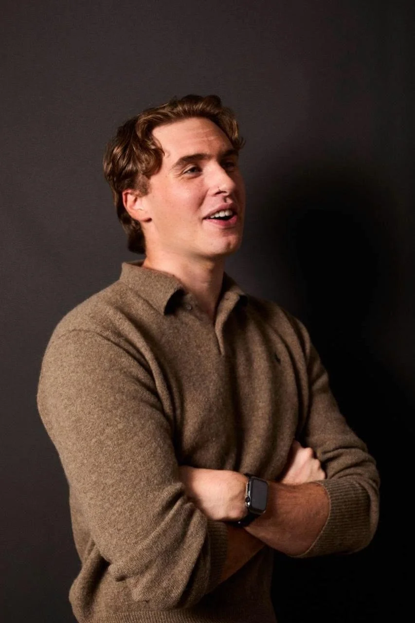 A young man with light brown hair, wearing a gray t-shirt, rests his face on his left hand with fingers touching his cheek, and has a nose ring.