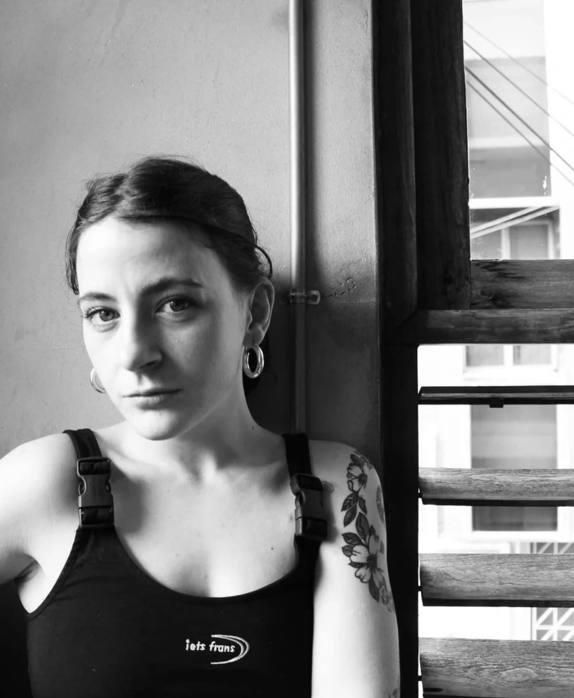 A woman with dark brown hair, wearing a gray t-shirt, hoop earrings, and a nose ring, standing outdoors near pink flowers and green foliage.