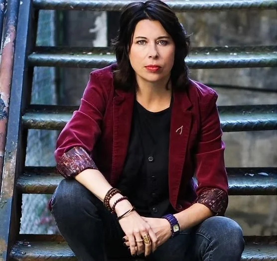 A woman with dark hair, wearing a burgundy blazer over a black shirt, sitting on metal stairs outdoors with a serious expression.