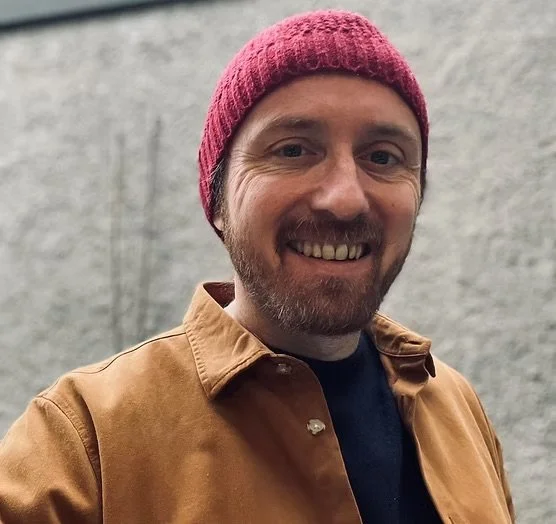 A man smiling, wearing a red knit beanie, a brown jacket, and a black shirt, standing outdoors against a gray textured wall.