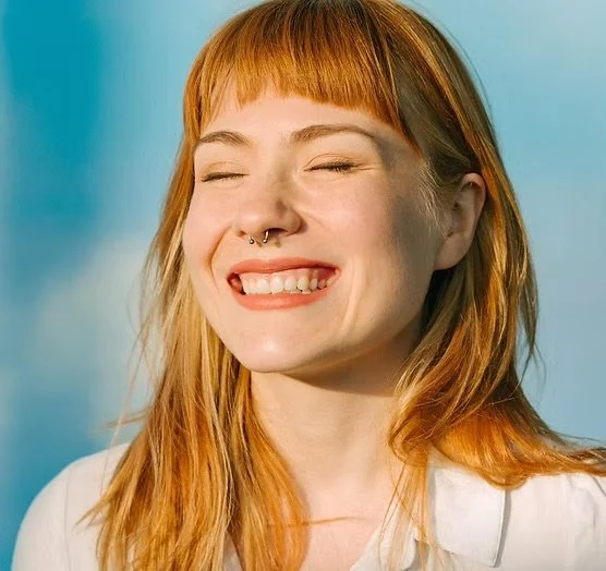 A young woman with red hair and a septum piercing smiling with eyes closed against a blue sky background.
