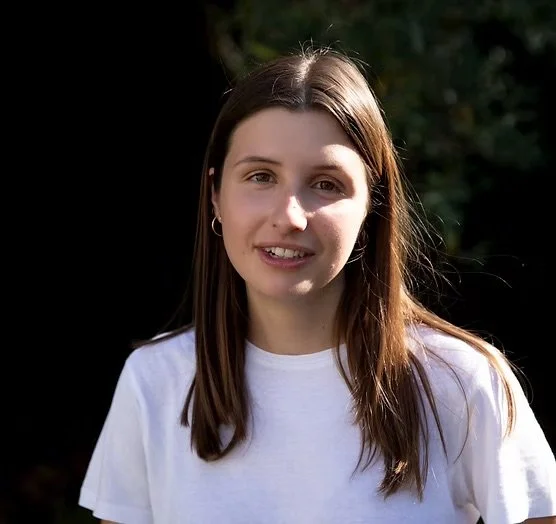 A young woman with long brown hair wearing a white t-shirt outdoors with green foliage in the background.