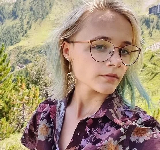 A young woman with short blonde hair and glasses taking a selfie outdoors in a lush green landscape with hills in the background.