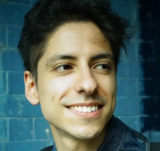 A young man smiling, wearing a dark shirt, standing in front of a blue brick wall.