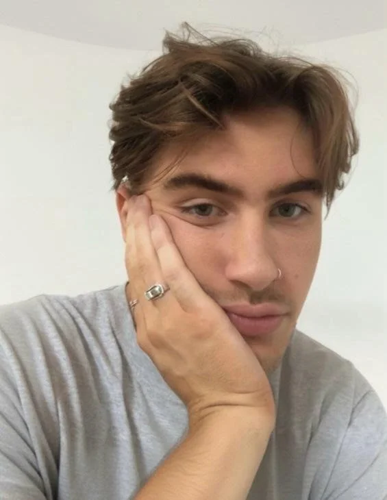 A young man with light brown hair, wearing a gray t-shirt, rests his face on his left hand with fingers touching his cheek, and has a nose ring.