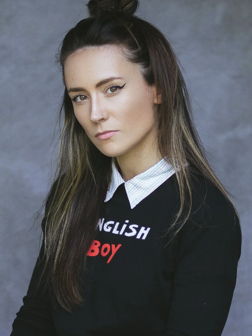 A woman with long wavy brown hair wearing a yellow and white gingham dress standing outdoors with green foliage in the background.