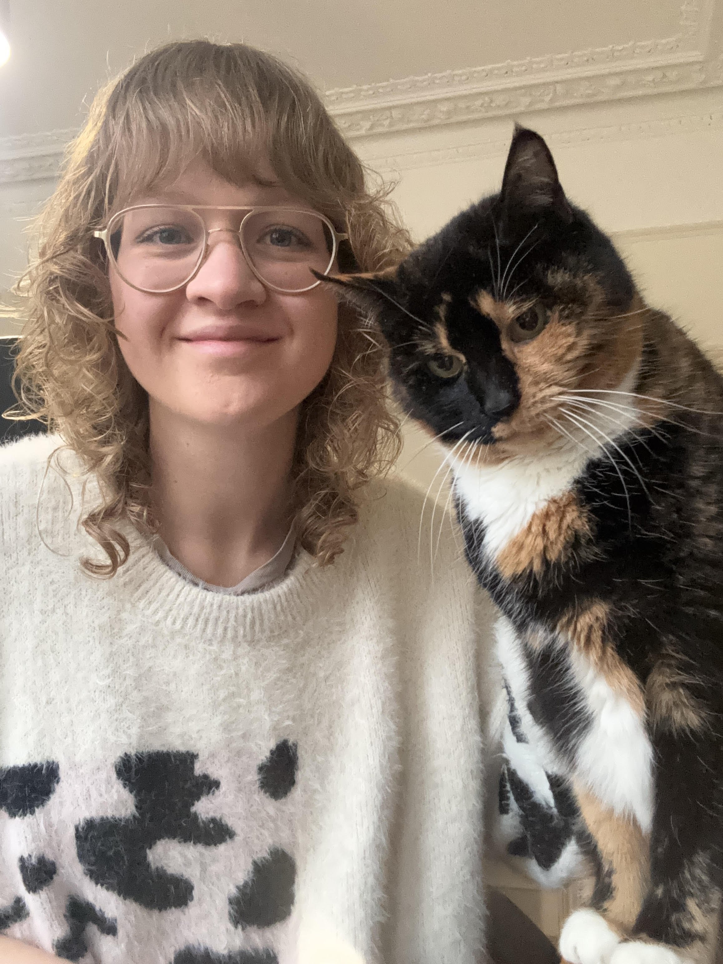 A woman with curly blonde hair and glasses smiling next to a calico cat.