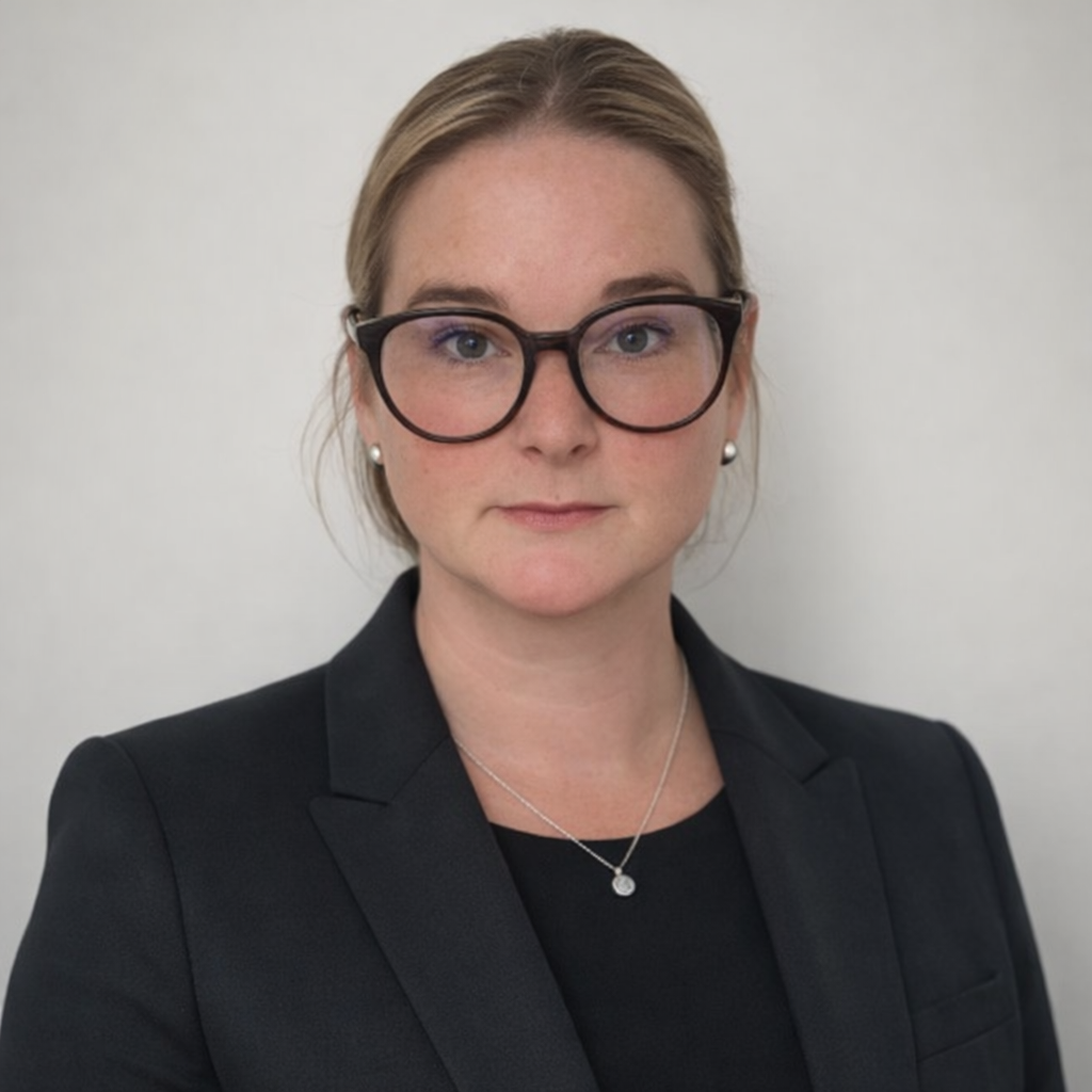 A woman with blonde hair, wearing glasses, pearl earrings, a black blazer, and a necklace, standing against a plain light-colored background.