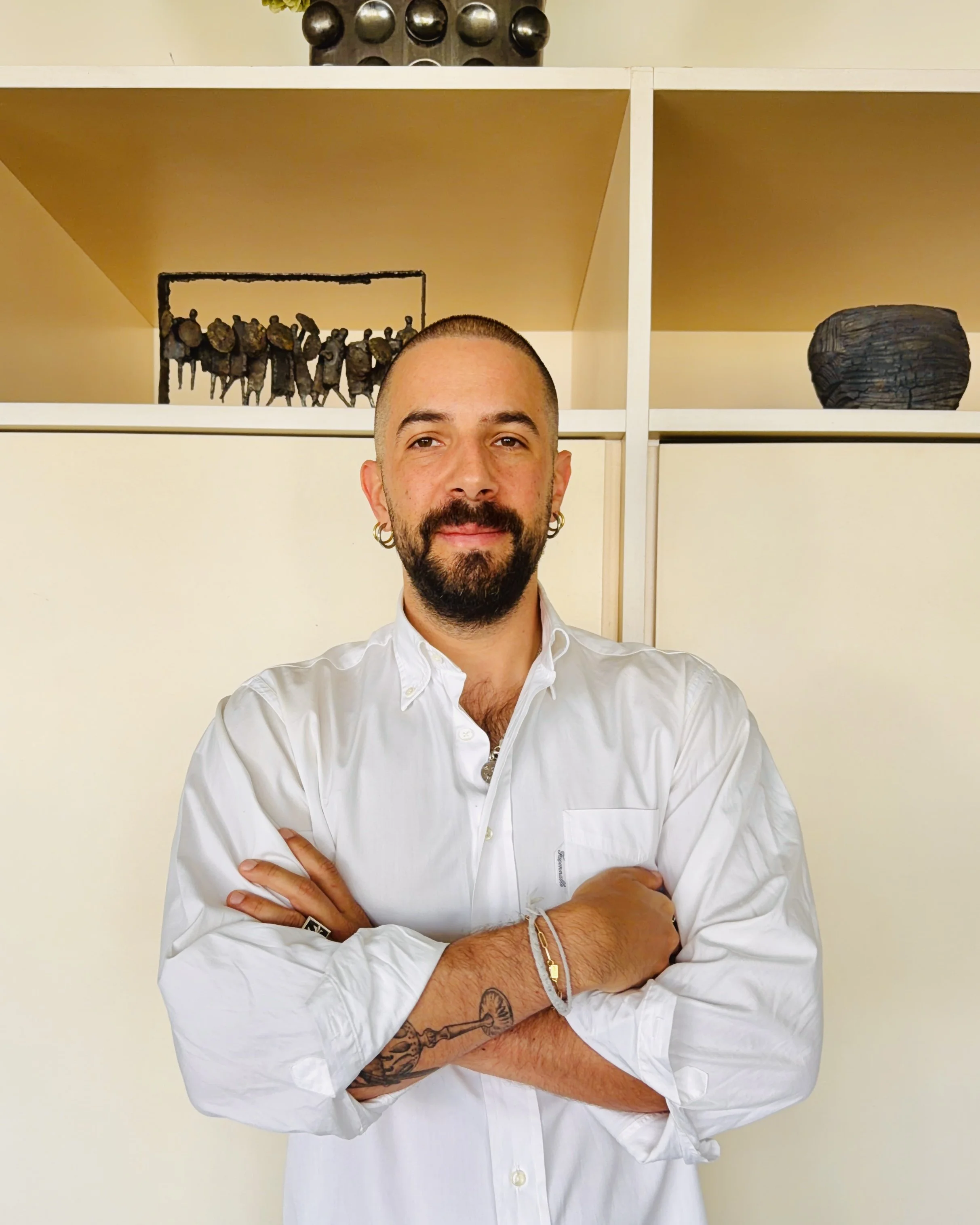 A man with a beard and short hair, wearing a white shirt, smiling with arms crossed, standing indoors in front of a yellow wall with art on shelves above.