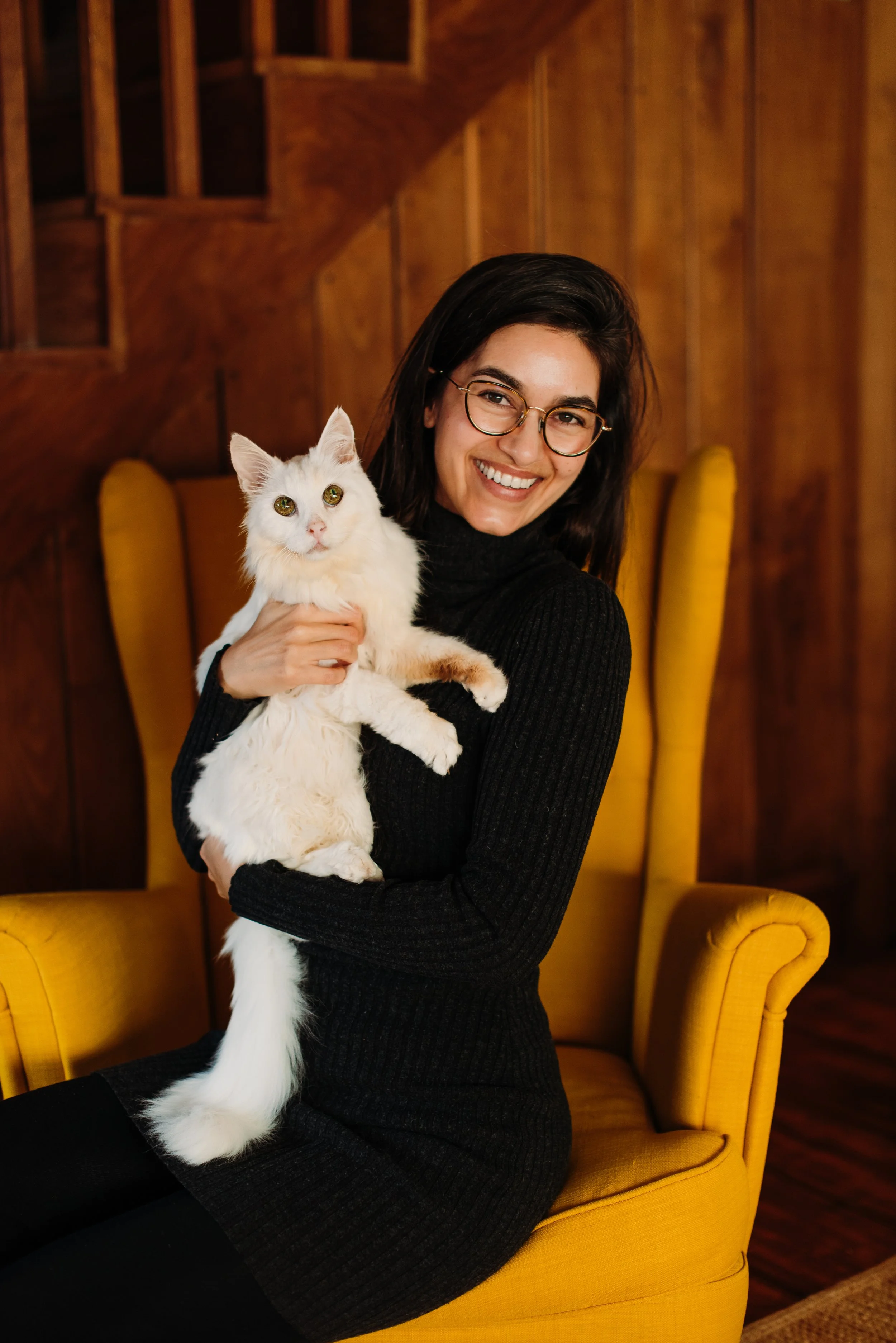 A woman with long dark hair, wearing glasses, a black jumper, sat on a yellow arm chair, holding a fluffy white cata necklace, standing against a plain light-colored background.