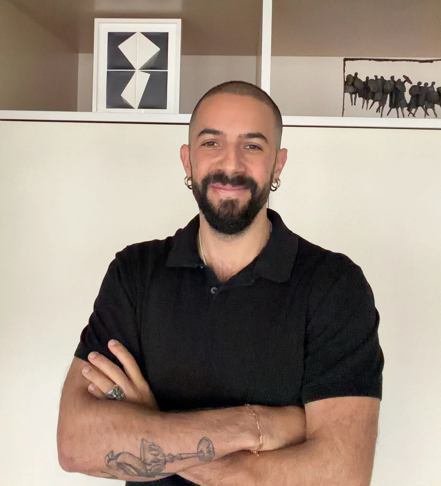 A man with a beard and short hair, wearing a black polo shirt, smiling with arms crossed, standing indoors in front of a white wall with art on shelves above.