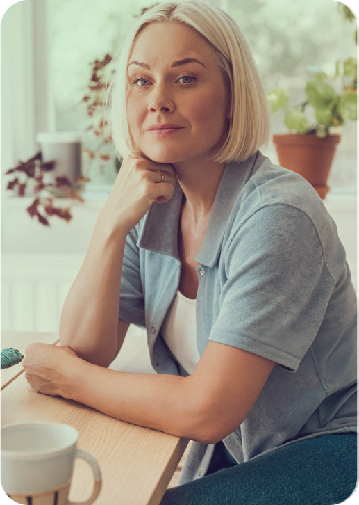 A woman with blonde hair, seated at a table in a bright room, resting her chin on her hand, wearing a light blue short-sleeved shirt, with plants and a window in the background.