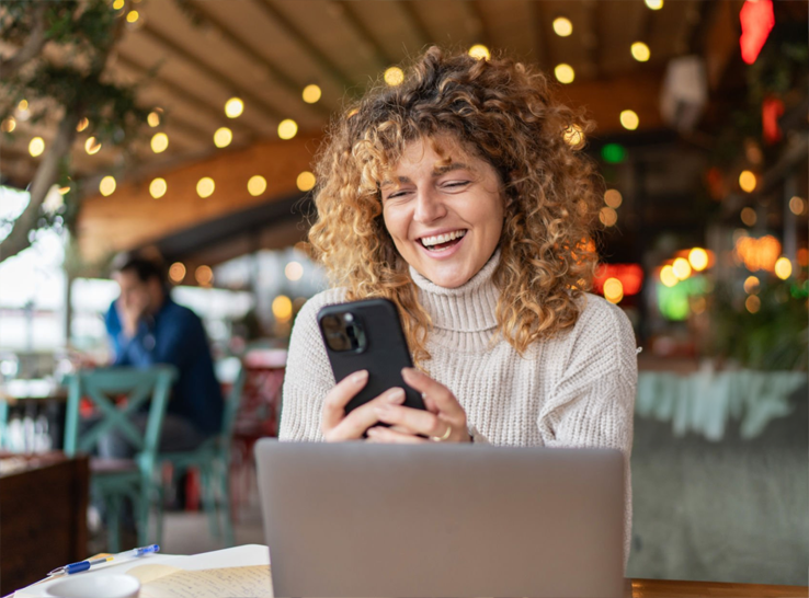 Woman with curly hair smiling and looking at her phone in a cozy, decorated cafe or restaurant.