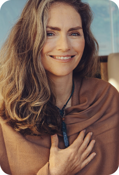 A woman with long, wavy brown hair smiling and holding her hand to her chest, wearing a brown sweater and a blue necklace, sitting indoors with a window in the background.