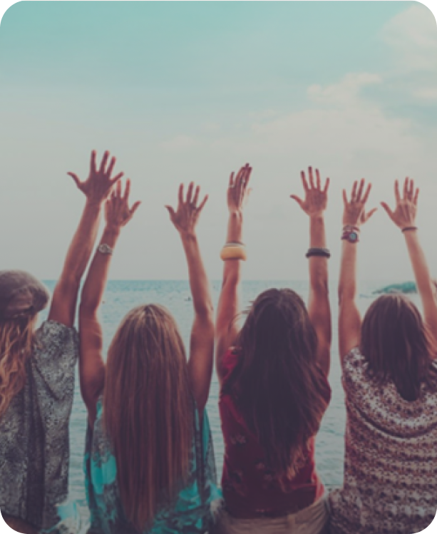 Four girls with long hair are sitting at the beach, facing the water, with their arms raised in the air during daytime.