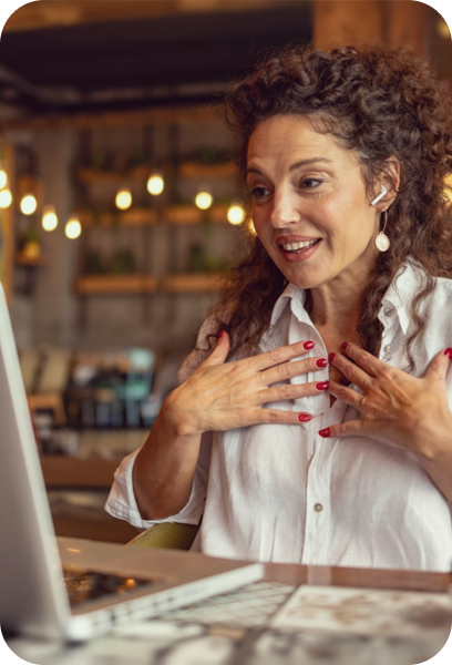A woman with curly hair wearing a white shirt and earrings is smiling and talking, possibly in a video call, in a cozy space with warm lighting and wooden shelves in the background.