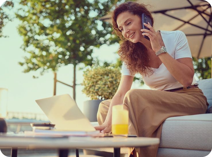 Woman sitting outside on sofa, talking on cell phone, with a laptop, papers, and a glass of orange juice on a table in front of her, in a sunny outdoor setting with trees and umbrella in the background.