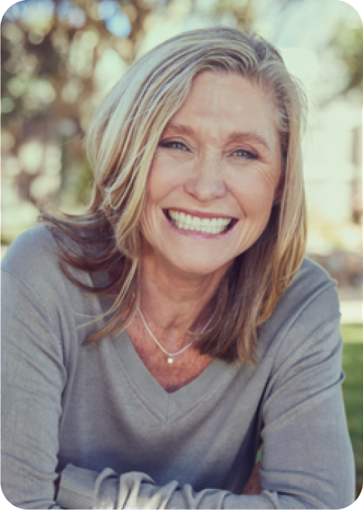 Smiling middle-aged woman with blonde hair, wearing a gray top and a necklace, outdoors with trees in the background.