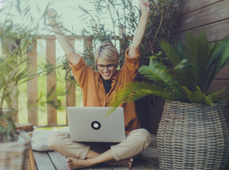Woman sitting cross-legged outside with a laptop, smiling and raising her arms in celebration, surrounded by potted plants.