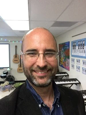 A man with glasses and a beard smiling, standing in a music classroom with guitars hanging on the wall and musical equipment including keyboards and a drum set.
