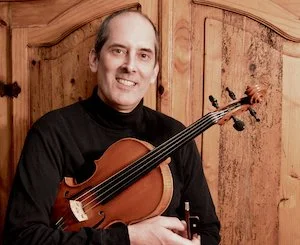 A man holding a violin and bow, smiling, in front of wooden cabinets.