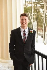 Young man in formal suit with boutonniere standing on porch with snow and trees outside