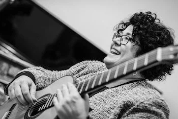 A woman with curly hair and glasses smiling while playing an acoustic guitar.