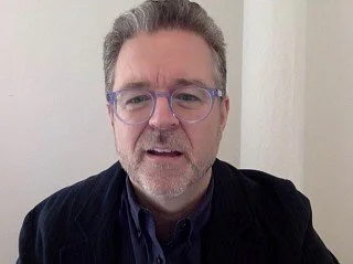 A middle-aged man with glasses and a beard, wearing a dark blazer, sitting in front of a light-colored wall.