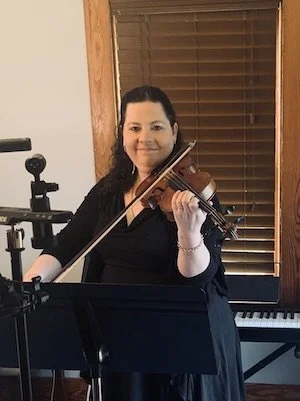 A woman playing the violin in a room with wooden blinds and a piano in the background.
