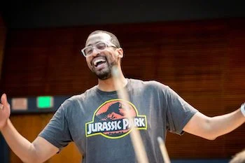 Man smiling and speaking, wearing a Jurassic Park t-shirt, in a room with wooden paneling.