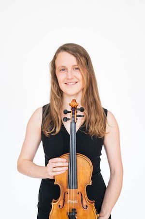 Woman holding a violin in front of her chest and smiling, wearing a black sleeveless top against a white background.