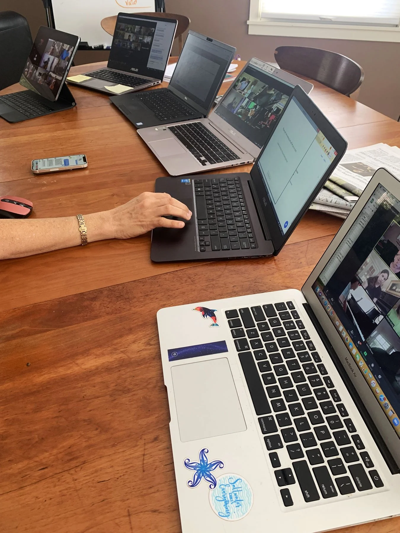 A conference table with multiple open laptops, a smartphone, a computer mouse, and scattered papers, with a person's hand operating one of the laptops.