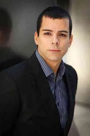 A man with short dark hair wearing a black blazer and a blue striped shirt, standing outdoors in front of a blurred background.
