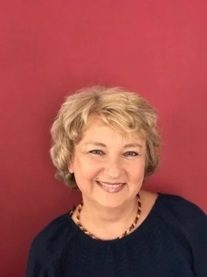 A smiling woman with short, curly blonde hair, wearing a black top and a beaded necklace, standing against a pink background.