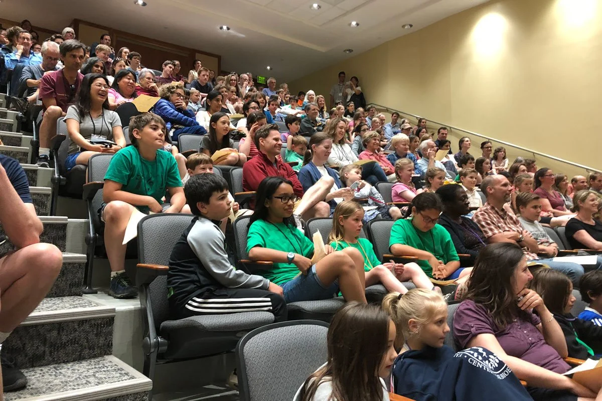 Audience seated in auditorium during a presentation or performance, diverse group of children and adults watching attentively.