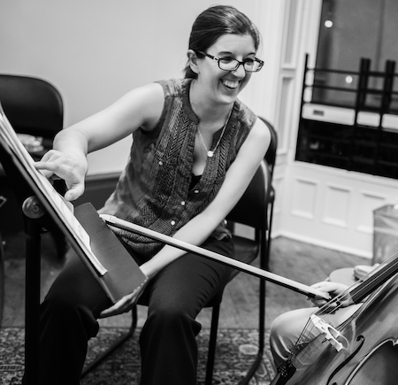 Woman playing the violin, smiling, in a room with chairs and a window.