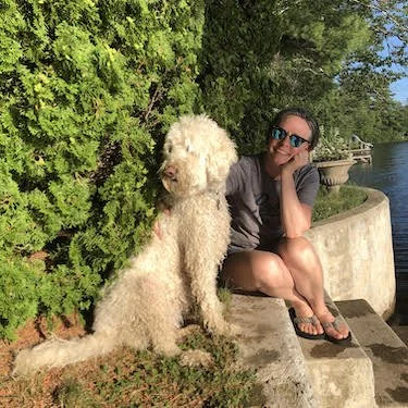 A woman sitting on a concrete ledge near water next to a large fluffy white dog, with green trees in the background.
