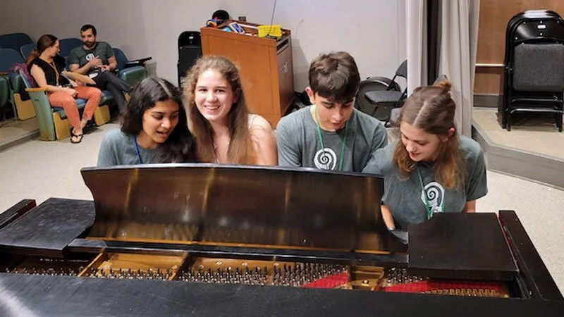 Four teenagers sitting at a piano, smiling and looking at the keys, with three people in the background sitting on chairs in a room.