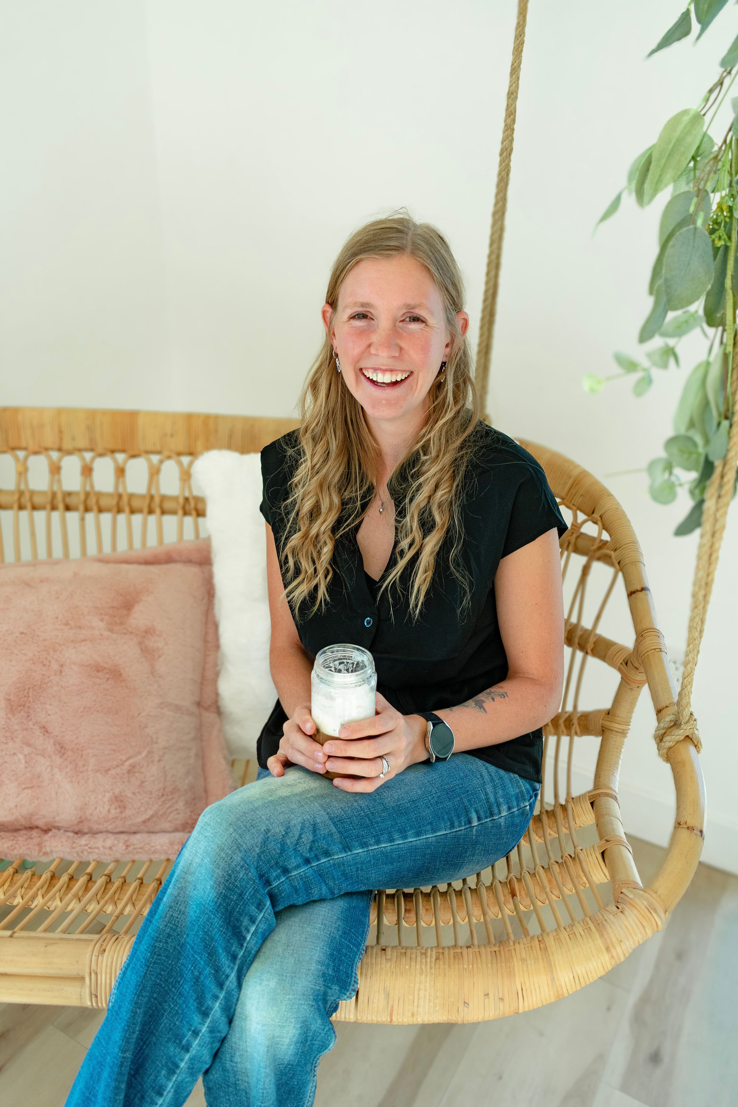 A young woman with long blond hair, smiling, sitting on a wicker bench indoors, holding a glass jar with a beverage, wearing a black top, blue jeans, and a smartwatch, next to a pink cushion and a white fluffy pillow, with green plants in the background.