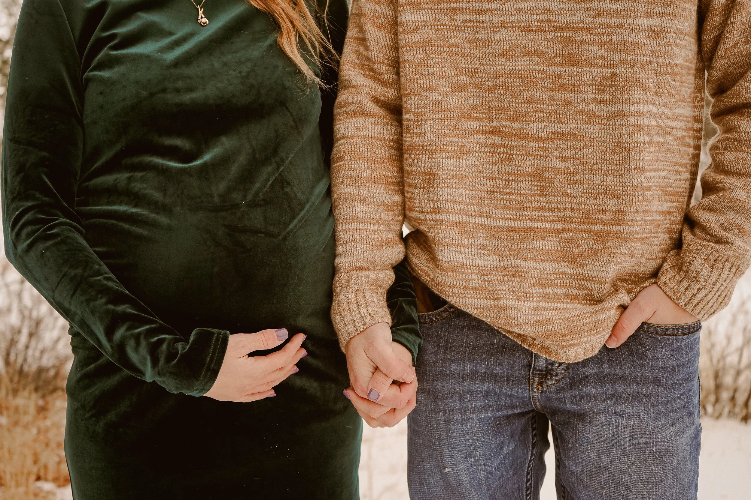 A pregnant woman and a man holding hands outdoors. The woman is wearing a dark green velvet dress and the man is dressed in a tan sweater and jeans.