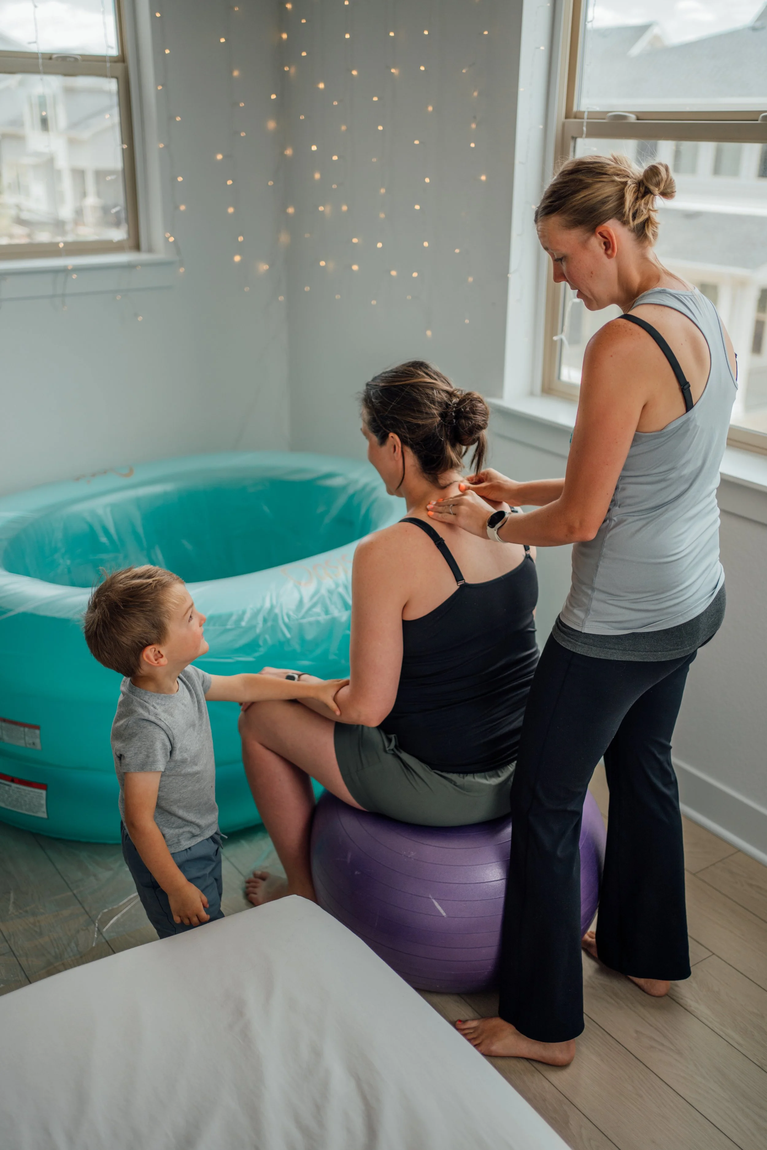 A pregnant woman seated on an exercise ball with a toddler holding her hand and a woman standing behind her giving a shoulder massage, near an inflatable pool in a sunlit room with string lights.