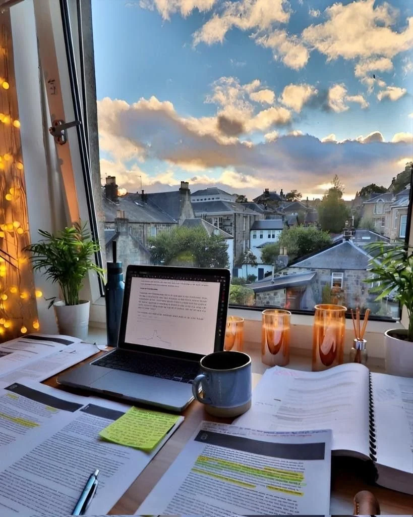 Estudio con vista a casas y cielo nublado, escritorio con computadora portátil, libros y documentos, tazas, plantas y luces decorativas.