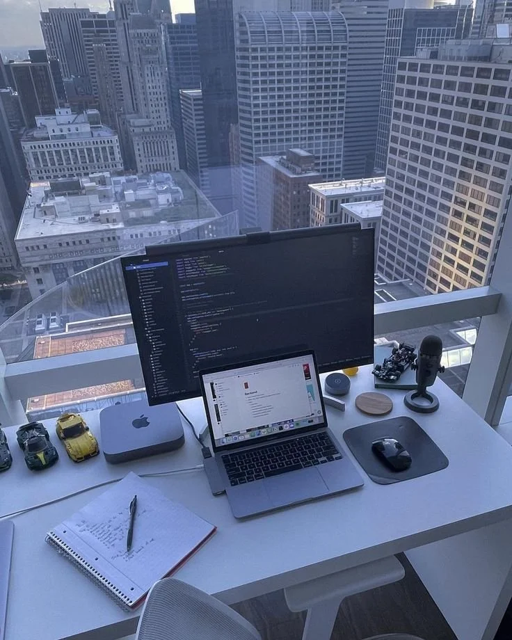 Mesa de trabajo en una oficina con vista a edificios altos, equipada con monitor, portátil, microfono, y objetos diversos.